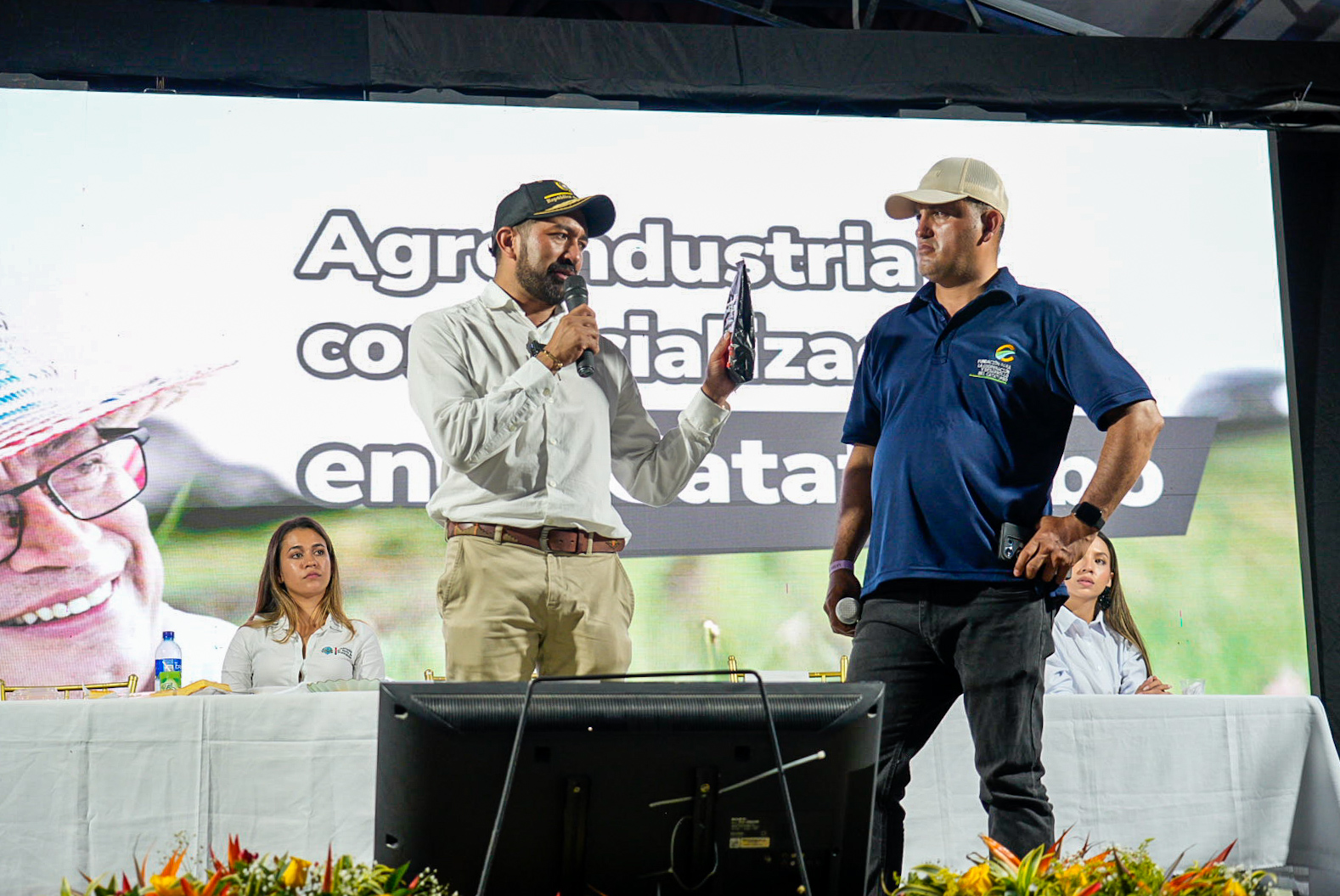 Presidente de la ADR sosteniendo un paquete en una mano y en la otra un micrófono y a su lado un hombre de camista azul